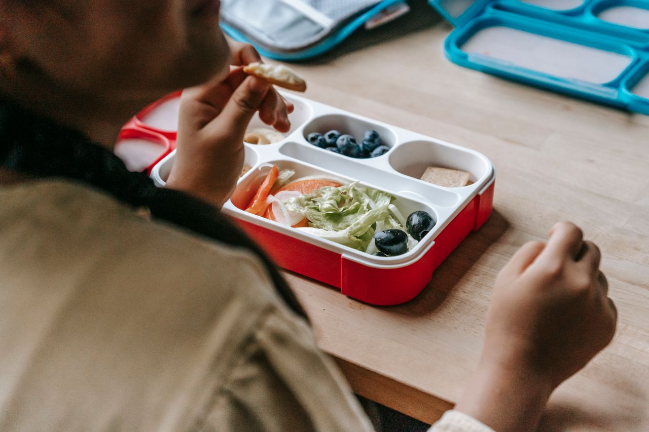Home Crop faceless teenage sitting at wooden table with lunch box full of healthy food